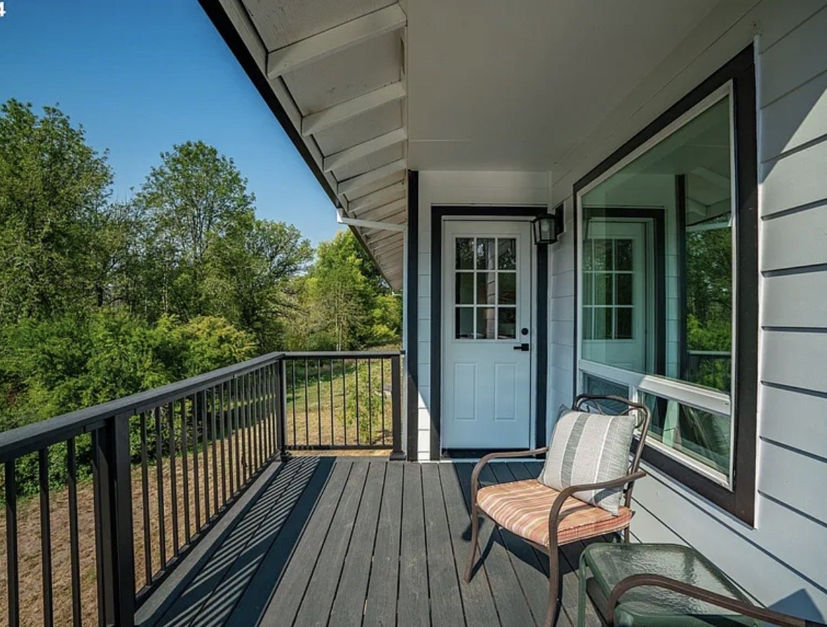 Balcony with a single chair, view of green trees, white house siding, door with glass panes, and railing on a sunny day.
