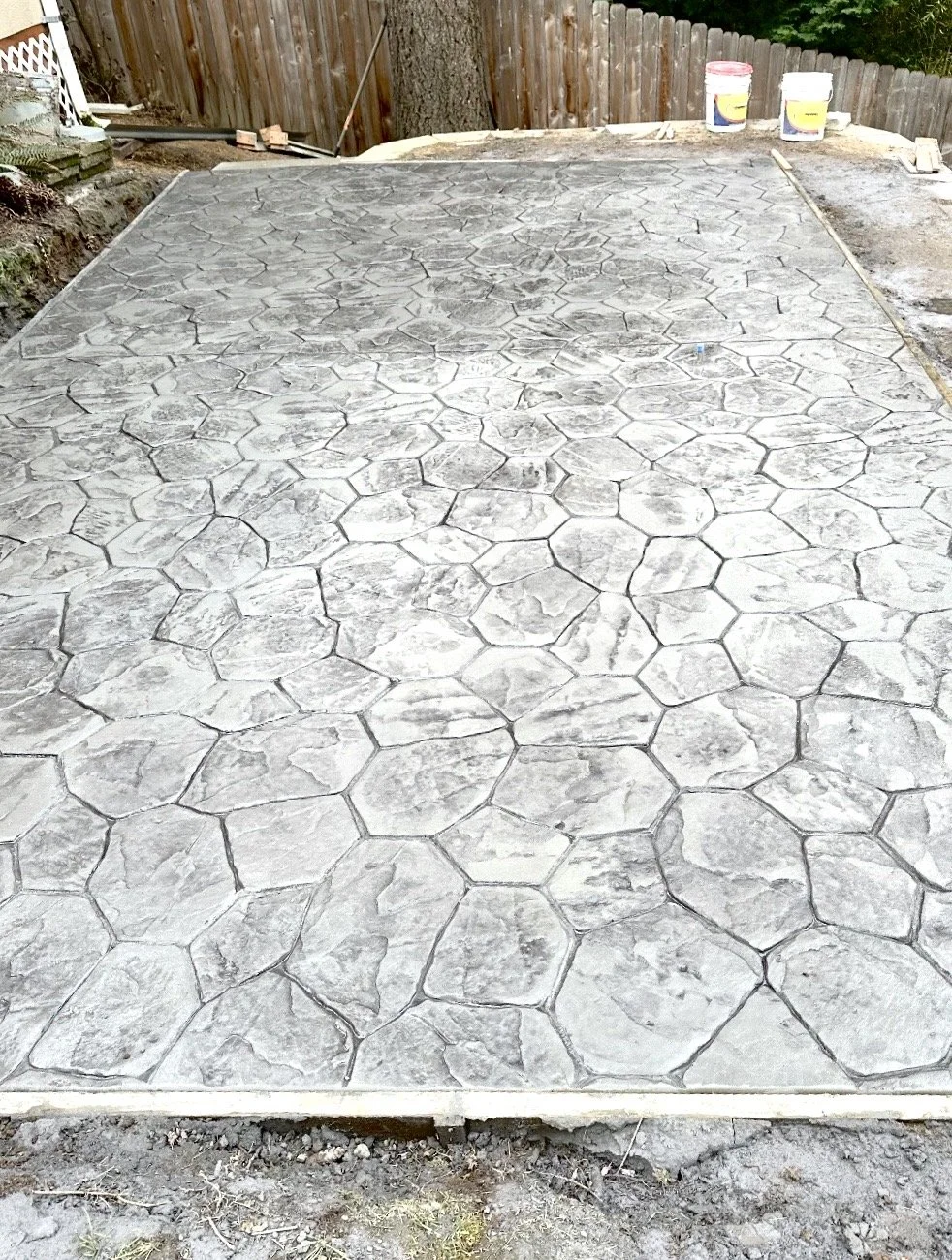 A newly poured concrete patio with a stamped pattern resembling hexagonal and irregular stones, surrounded by a wooden fence and construction tools in a backyard.