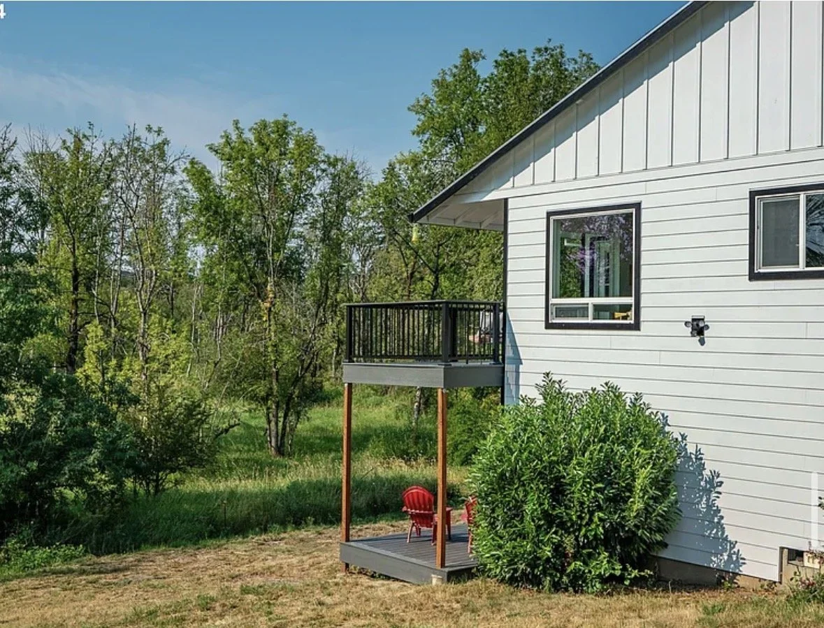 Backyard with a white house or shed, small deck with red chairs, green bushes, and trees in the background under a blue sky.