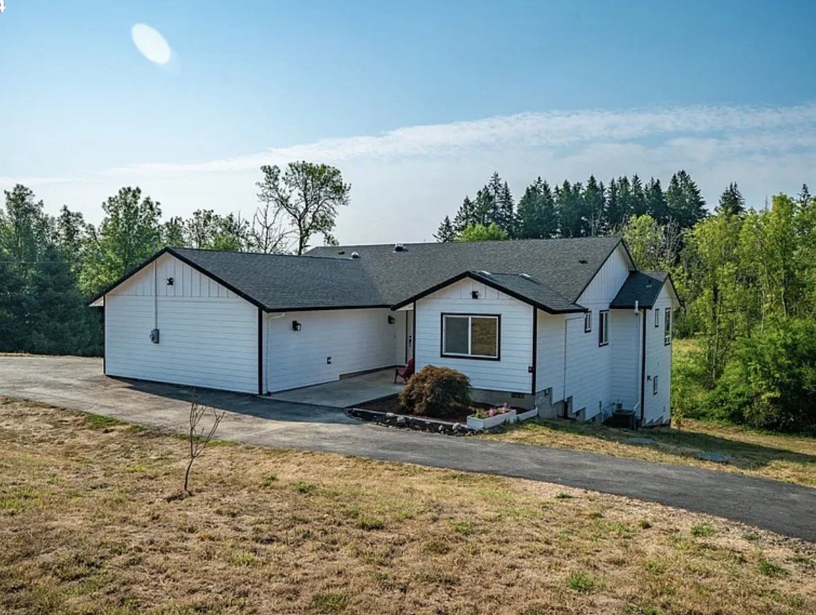 A white house with black trim, situated on a sloped yard with a paved driveway, surrounded by trees under a blue sky.