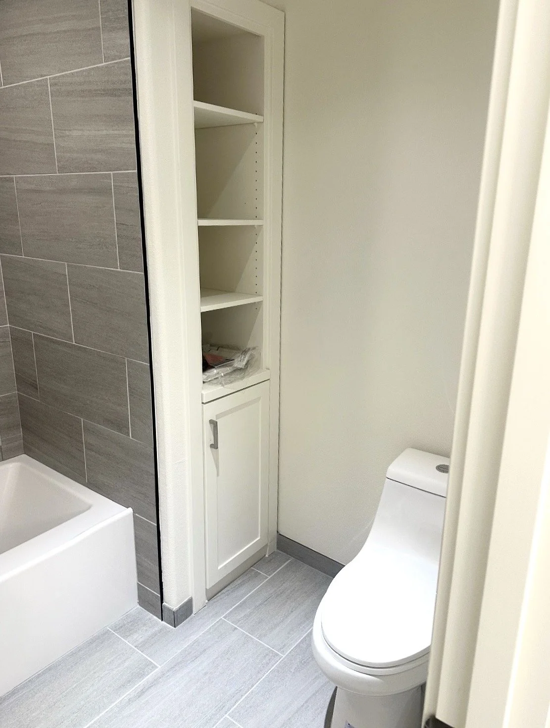 Bathroom with a white toilet, a built-in white cabinet with open shelves, and a section of a bathtub with gray tiles on the wall.