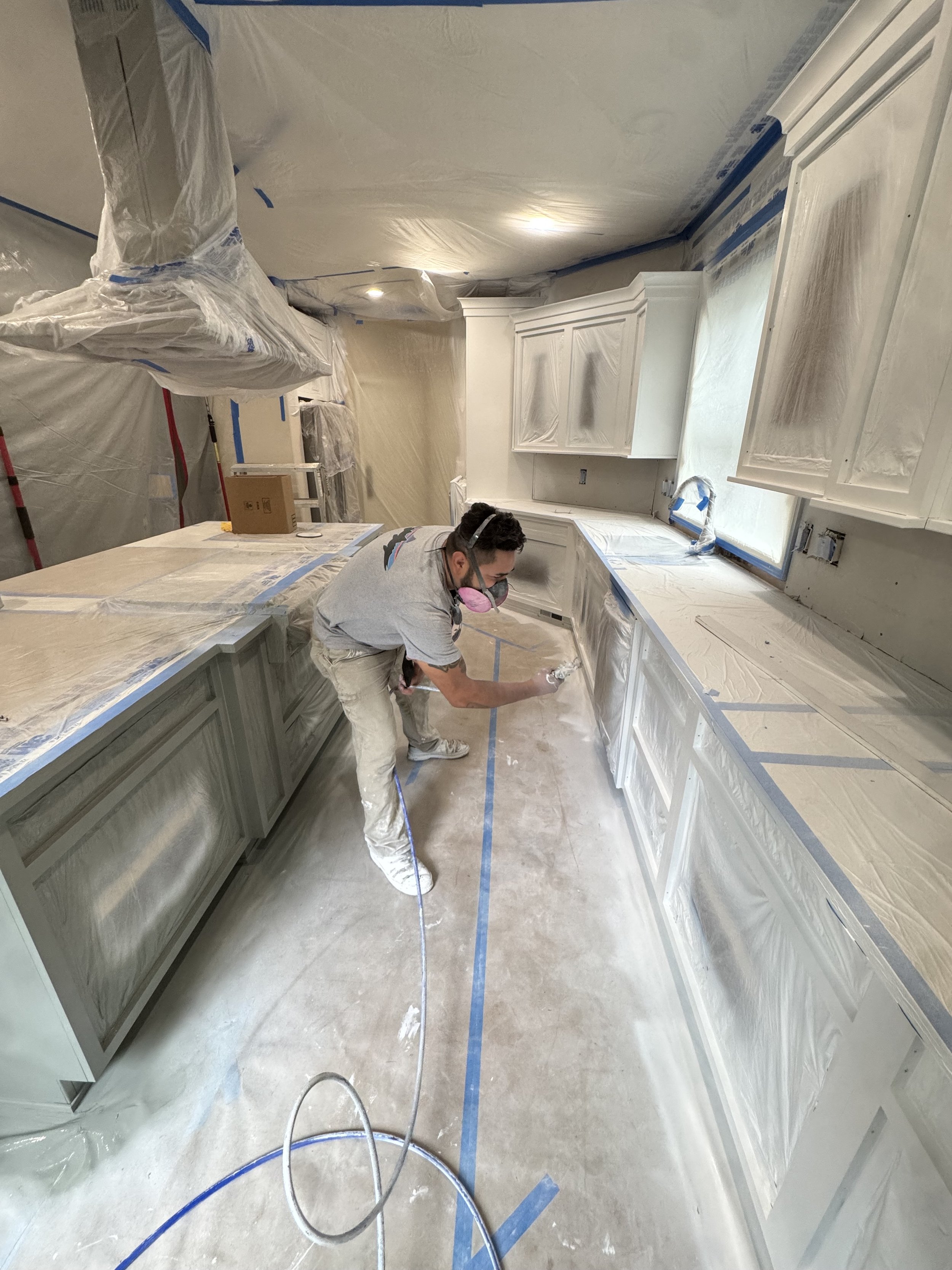 A person wearing a mask and protective glasses painting cabinets in a kitchen under renovation, with plastic coverings and taped surfaces.