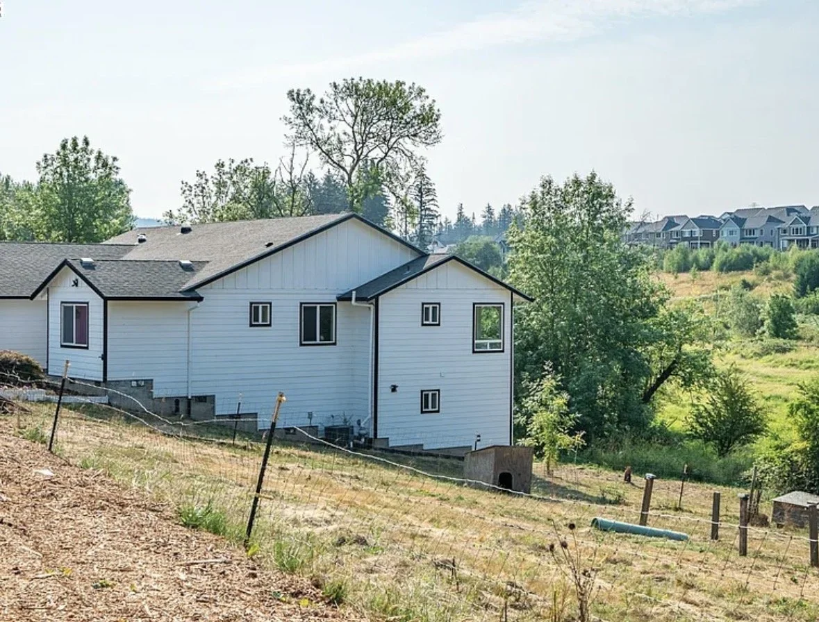 White house on a sloped grassy hill with trees and other houses in the background.