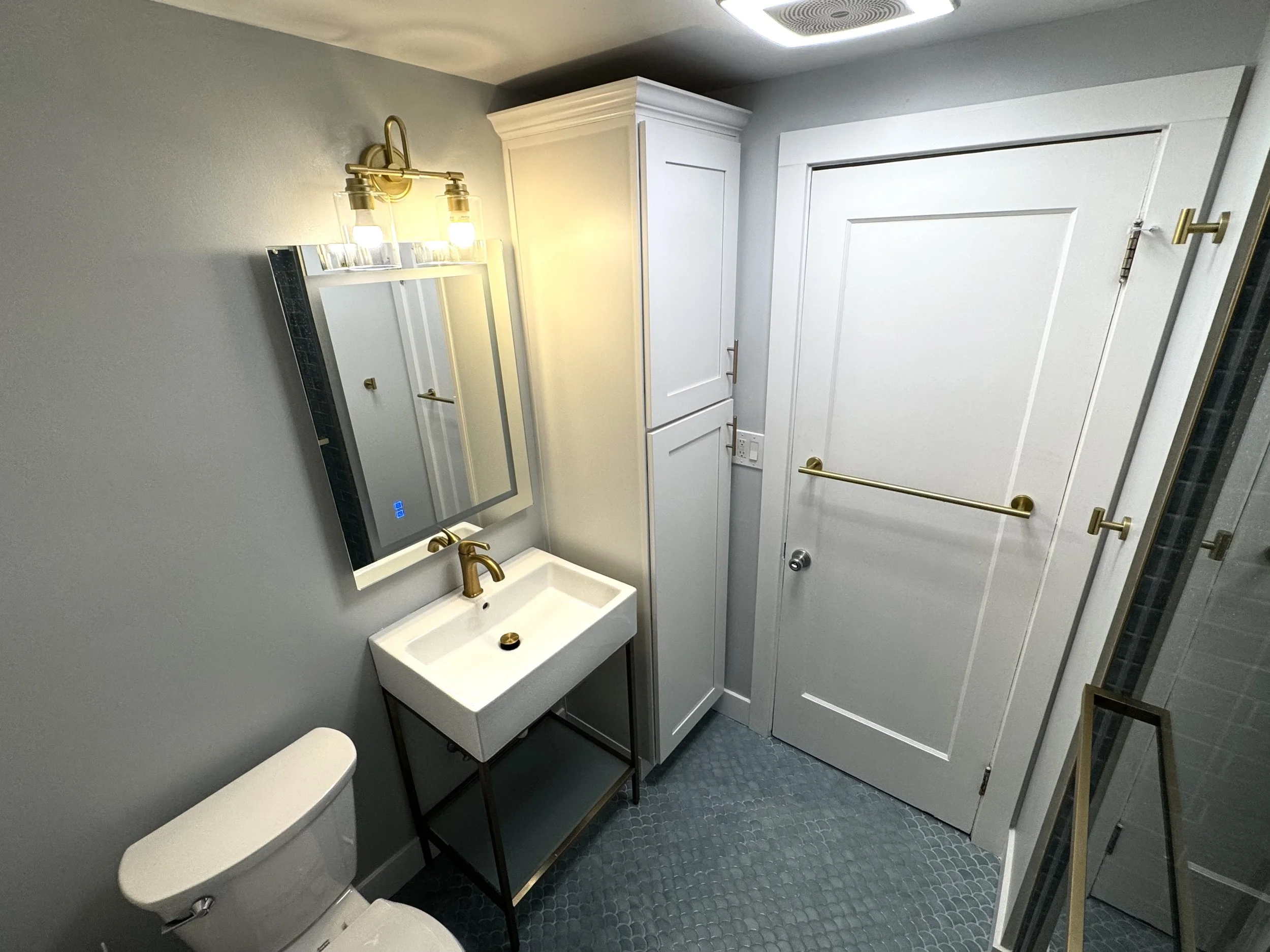 Modern bathroom with gray walls, a white vanity with a gold faucet, wall-mounted mirror, gold light fixture, white cabinet, white door with a gold towel bar, and glass shower door.