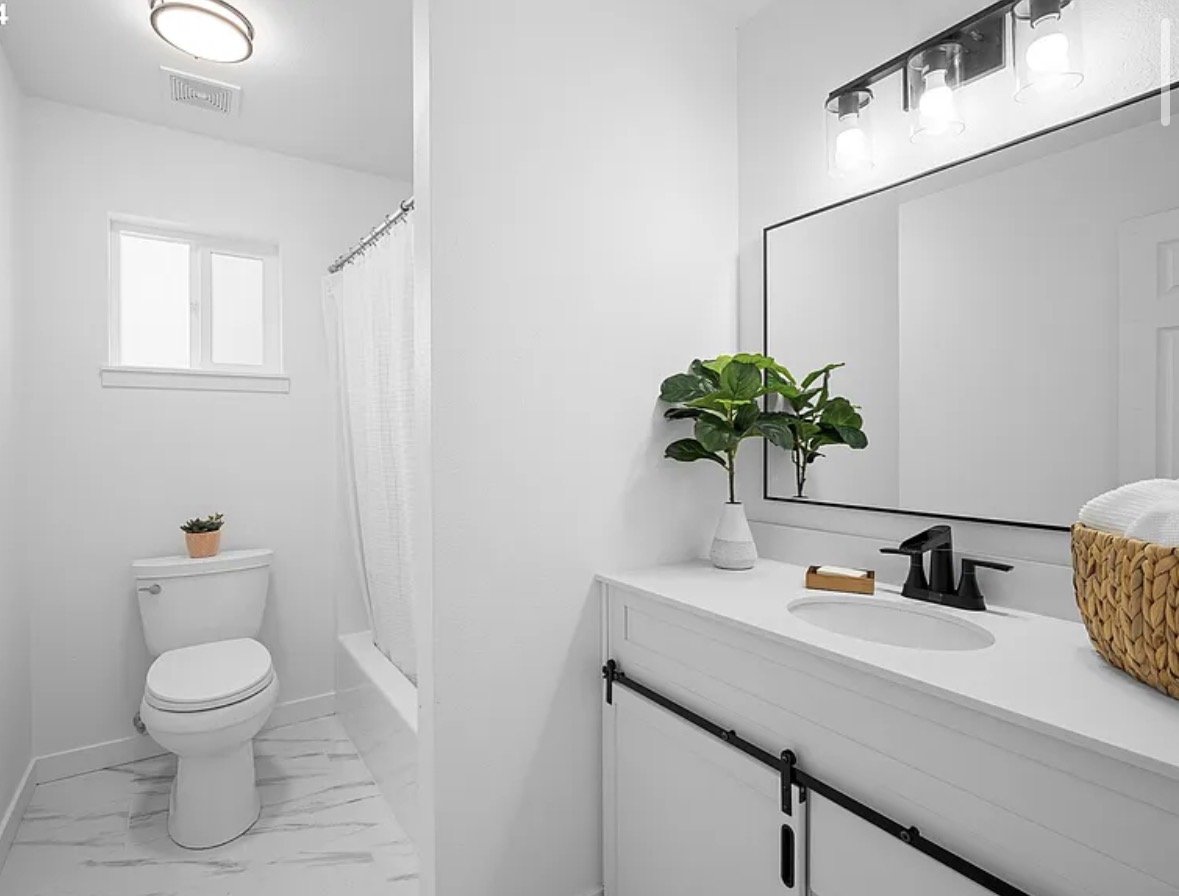 A minimalist bathroom with white walls and fixtures, a small window, a toilet with a potted plant on top, a shower with a white curtain, a white vanity with a sink, a large mirror, a black faucet, a potted plant, a soap dish, and a wicker basket on the counter.