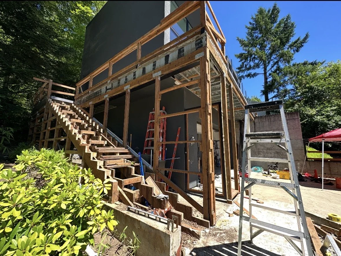 Construction site of a building with wooden framing, stairs, and scaffolding, surrounded by trees on a sunny day.