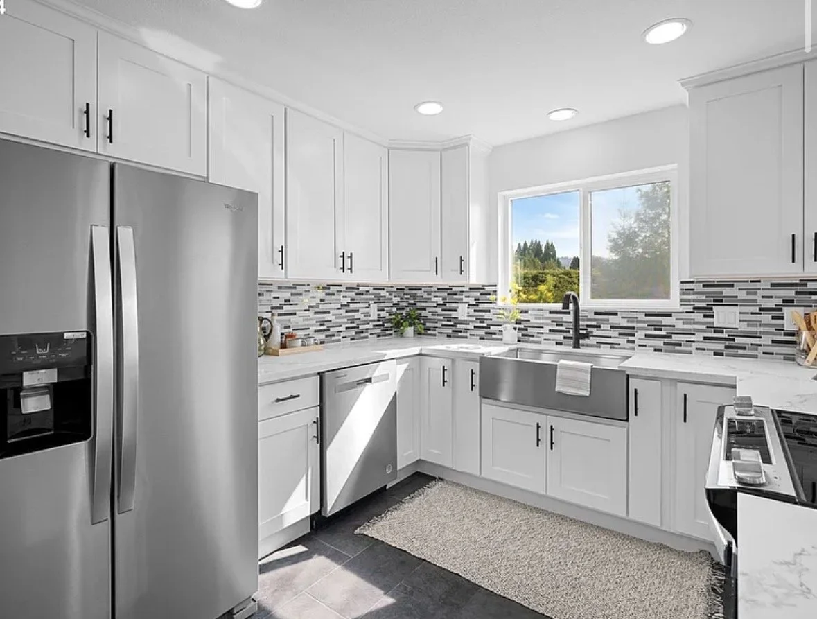 Modern kitchen with white cabinets, a gray and black backsplash, stainless steel appliances, and a window with a view of a green landscape.