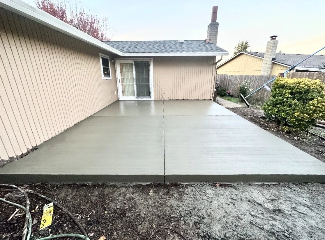 Newly poured concrete patio with a neutral-colored house and sliding glass door, surrounded by a yard with a garden bed, shrub, and neighboring houses.