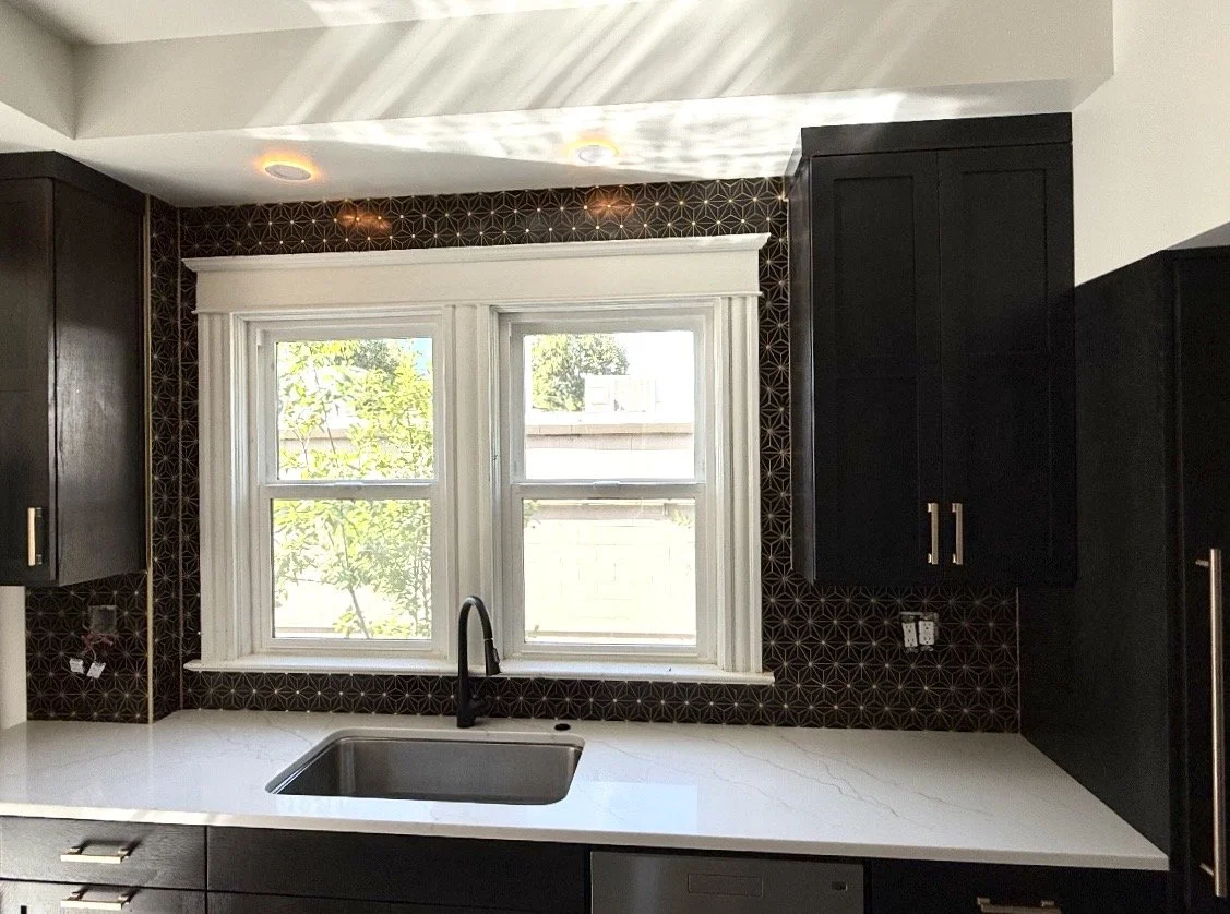 Kitchen with a double window, dark cabinets, white marble countertop, black faucet, and geometric patterned black backsplash.