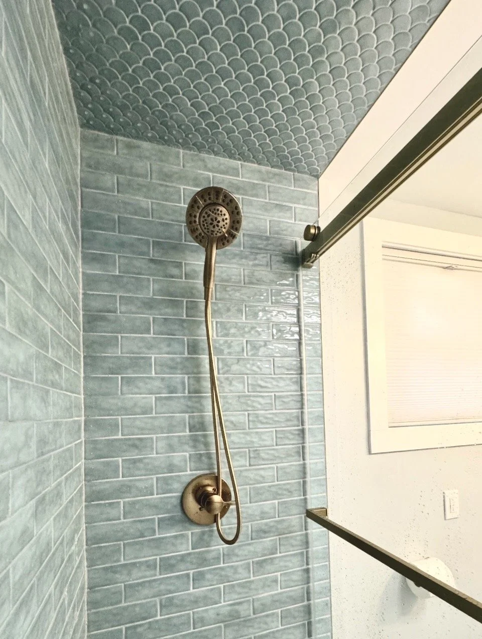 Bronze showerhead and faucet against light blue brick tile in a bathroom shower with a glass sliding door and a mirror.