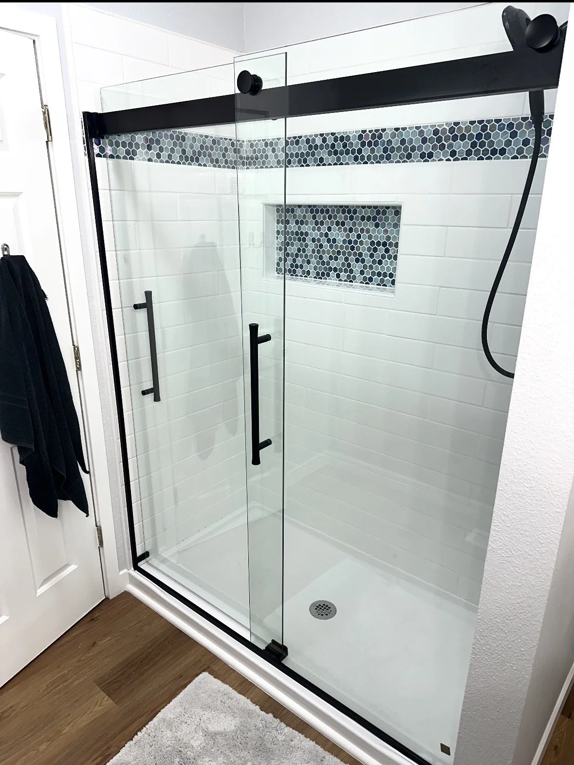 A modern walk-in shower with glass doors, black handles, a hexagon tile accent strip, a built-in shelf, and a black showerhead.