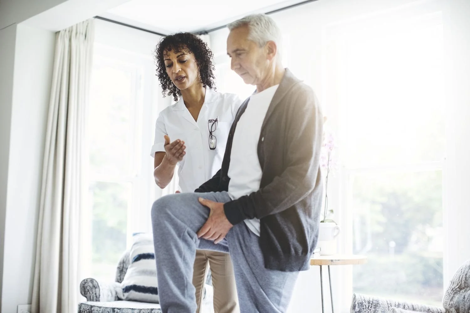 A healthcare professional, possibly a physical therapist or doctor, assisting an elderly man with knee pain by holding his knee and thigh while he balances on one leg in a well-lit room with large windows, curtains, and a floral armchair.