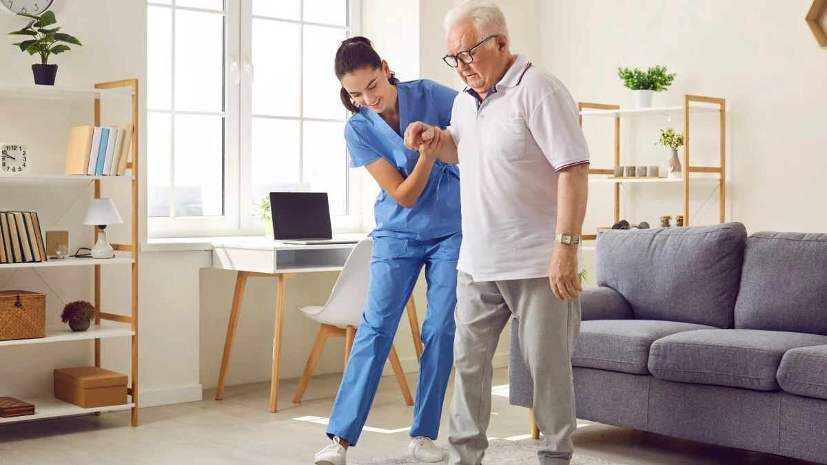 Nurse assisting an elderly man using a walker in a bright living room.