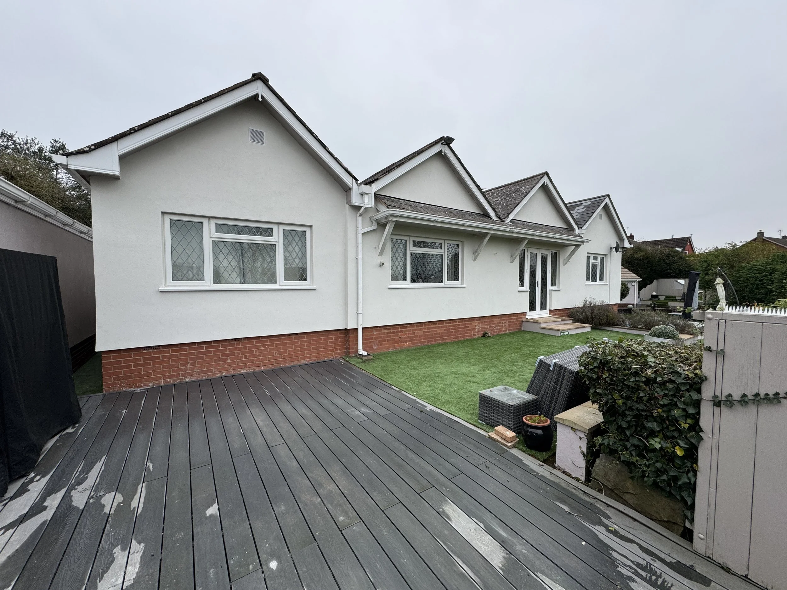 Front view of a white house with brick foundation and gabled roof, with a deck in the foreground and a lawn with garden furniture.