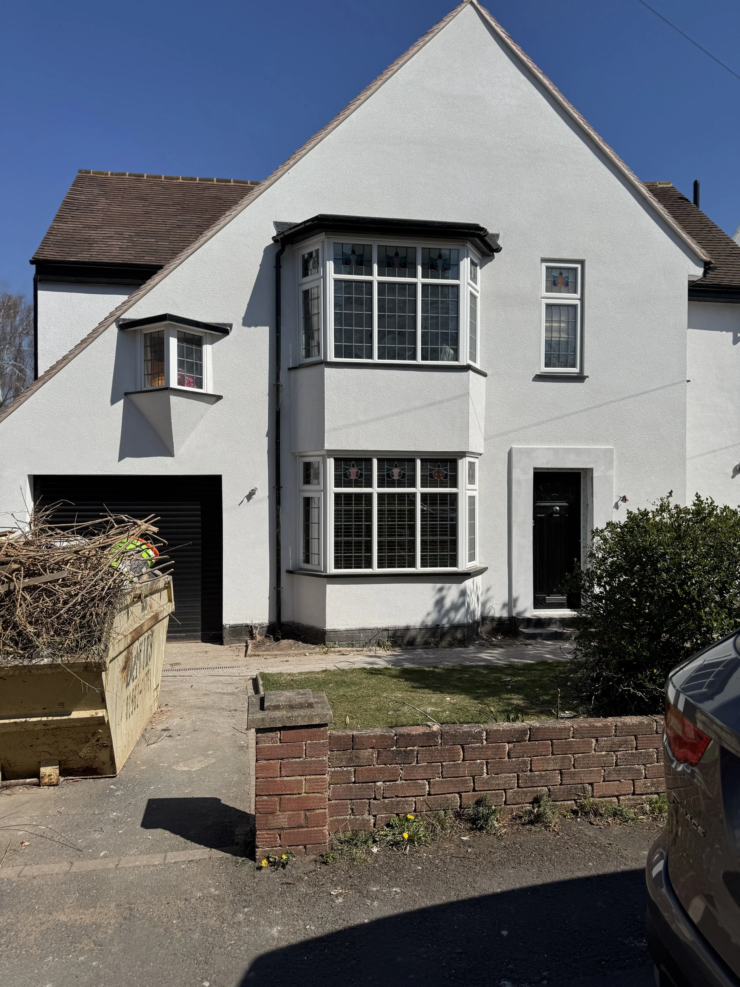 A white two-story house with a sloped roof, black front door, and large bay windows with stained glass accents. There is a small garden area in front with a low brick wall, bushes, and a driveway with a car parked partially visible.