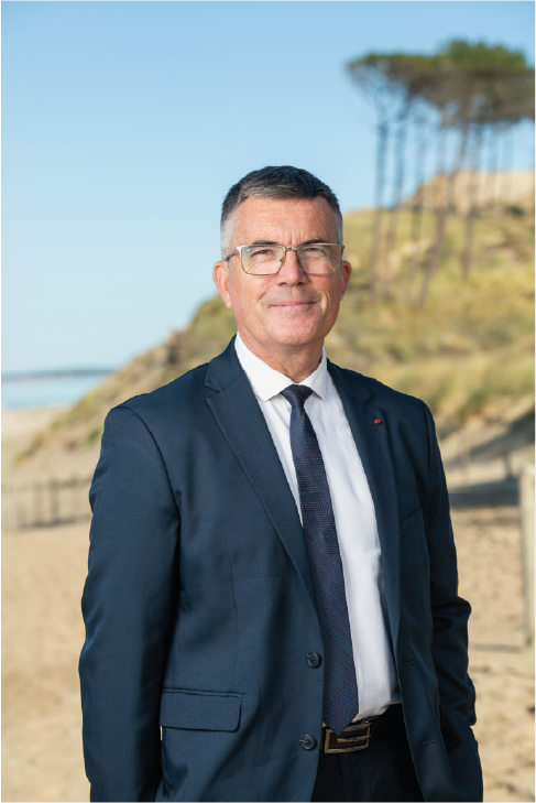 Un homme en costume bleu portant des lunettes, debout sur une plage avec des collines et des arbres en arrière-plan.