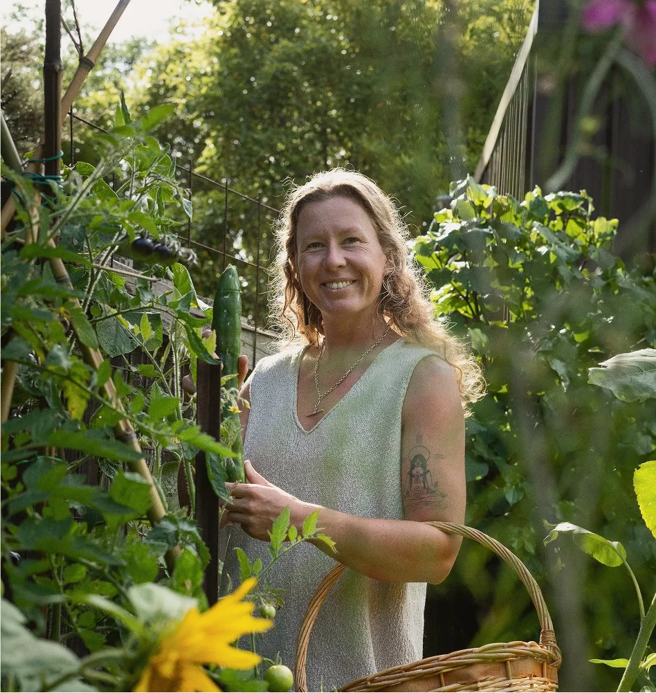 A woman with wavy hair smiling in a garden, holding a zucchini, surrounded by lush green plants with a basket hanging from her arm.