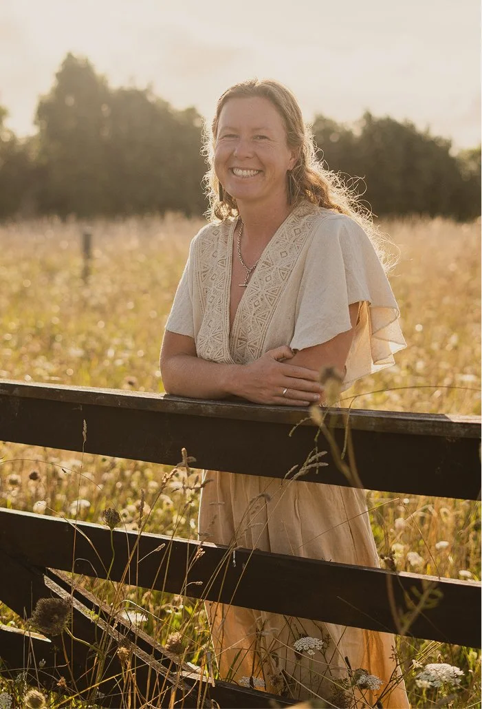 Smiling woman with wavy hair leaning on a wooden fence in a sunlit field with tall grass and wildflowers.