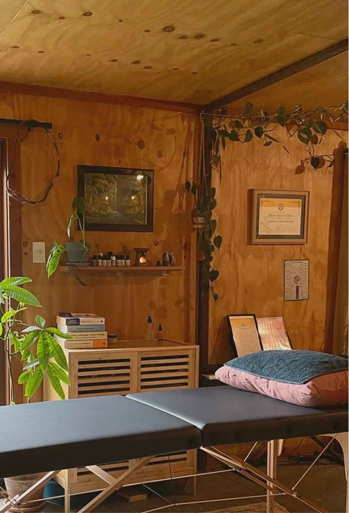 A massage therapy room with a massage table covered with a dark sheet, and a pillow at the head of the table. The room has wooden walls and ceiling with indoor plants, framed pictures, and certificates on the walls. There is a small cabinet with books and essential oils or bottles on it, and a wooden shelf with decorative objects.