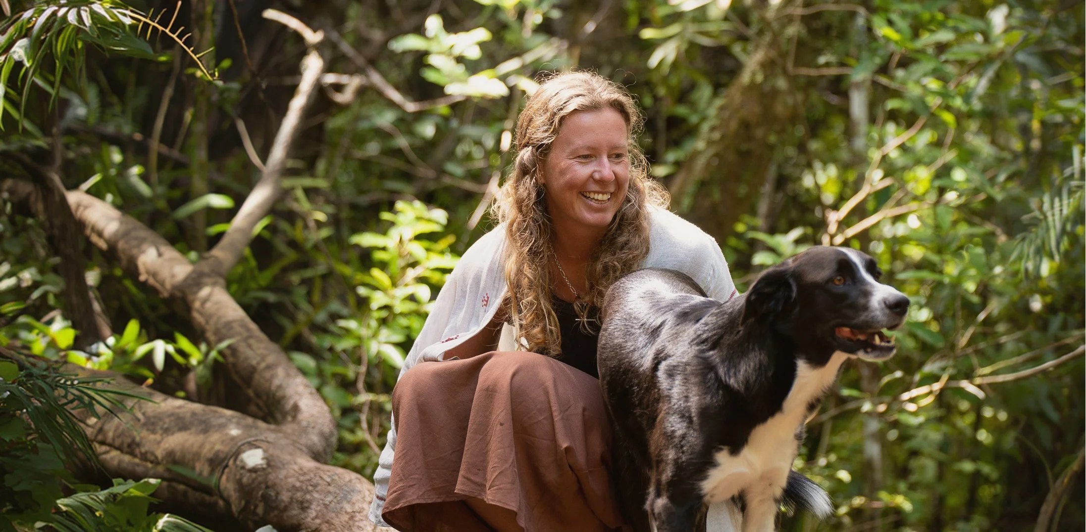 A woman with long curly hair, wearing a white cardigan and brown pants, is smiling while crouching beside a black and white dog in a lush, green forest.
