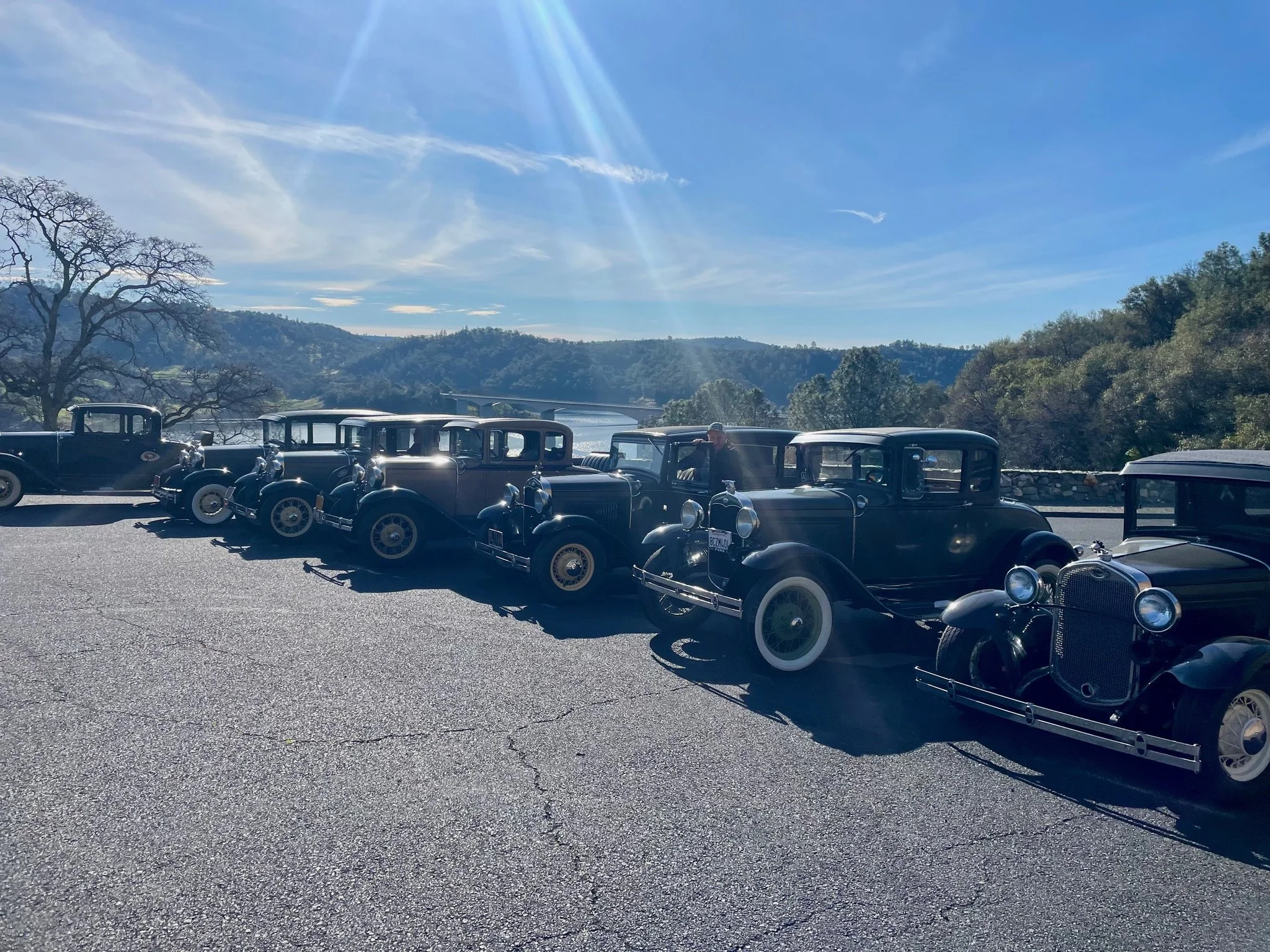 Line of vintage black cars parked on a paved area with a scenic background of hills, trees, and a river, under a blue sky with sunlight.