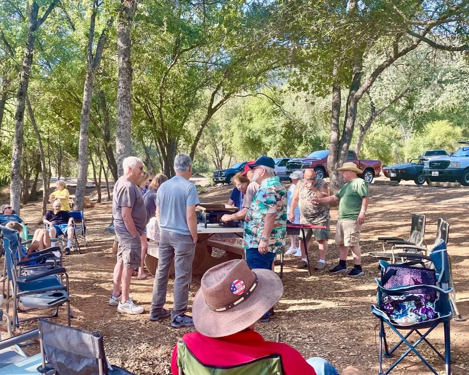 People gathered outdoors under trees, some sitting in chairs and others standing around a picnic table, with vehicles parked in the background.