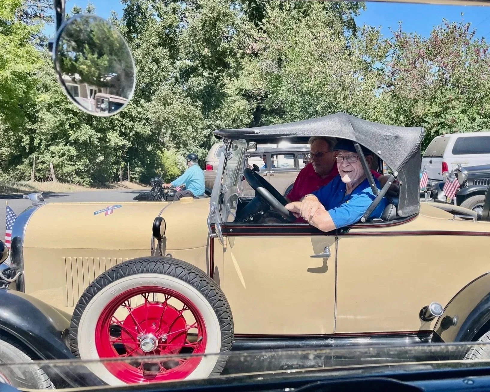 A vintage beige car with red wheels being driven by two elderly men, one wearing a blue shirt and sunglasses, and the other in a red shirt, during a parade. The car has small American flags attached to the rear. A motorcyclist rides in the background, and trees with green leaves surround the scene under a clear blue sky.