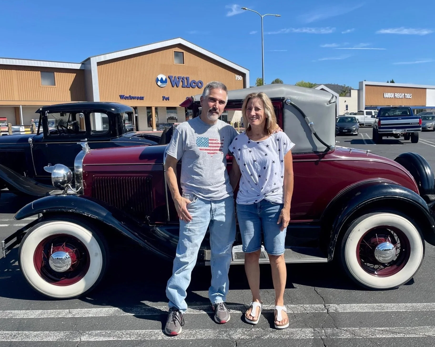 A man and woman standing together in a parking lot, smiling, with a vintage red and black convertible car behind them. The background features a Wilco hardware store and a row of parked cars under a blue sky.