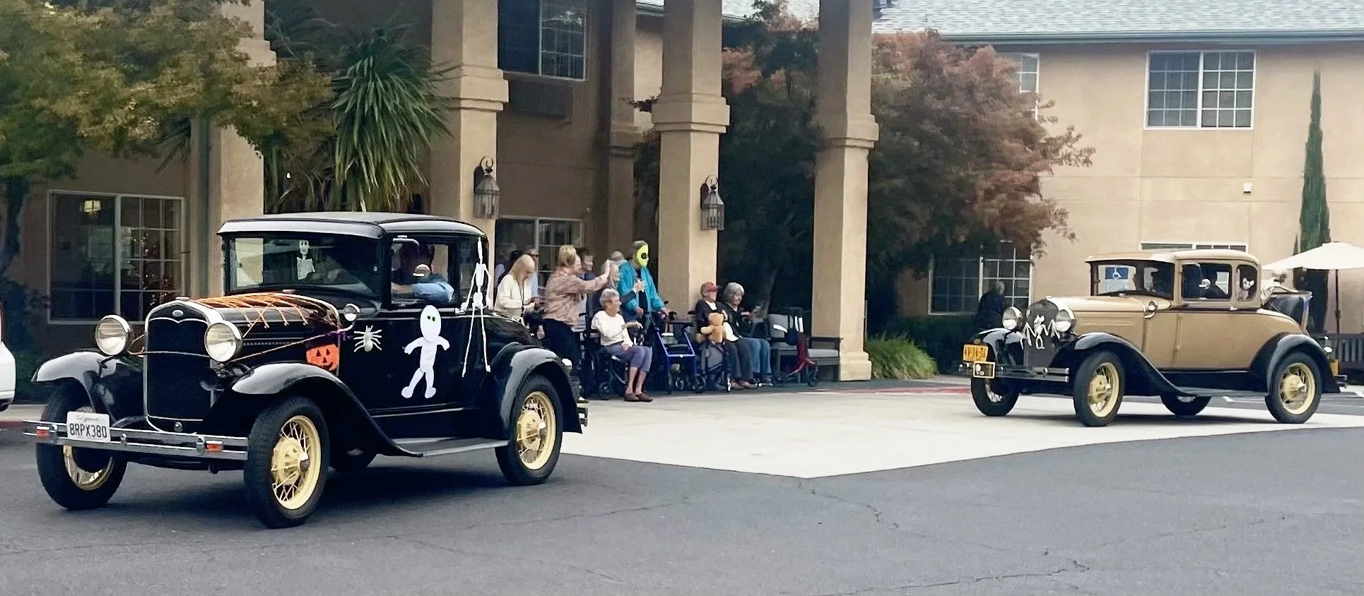 Two vintage cars parked outside a building with people, some in wheelchairs, sitting on the porch watching the scene.