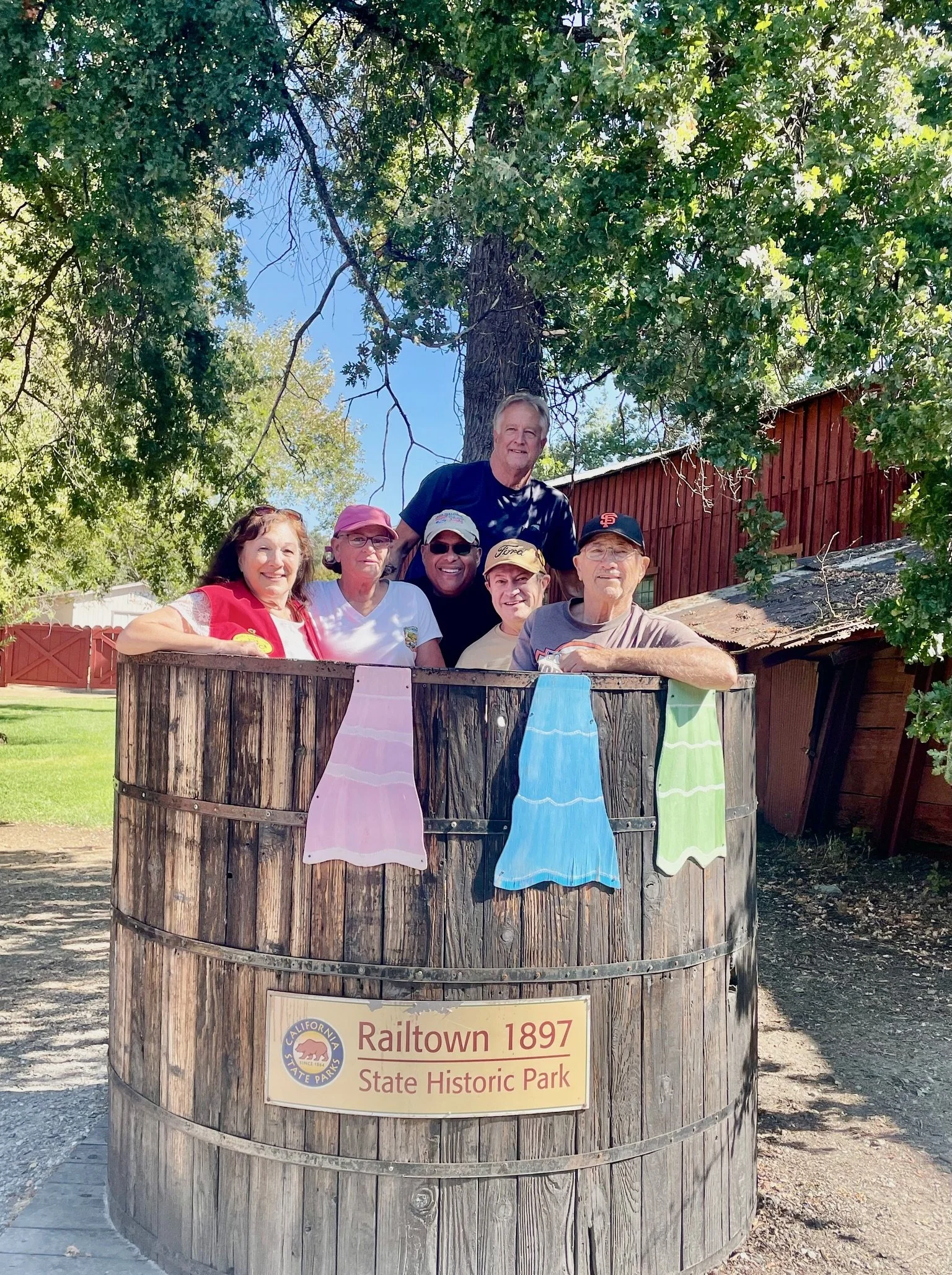 A group of seven people posing inside a wooden tub at Railtown 1897 State Historic Park, with colorful towels hanging on the side, under a large green tree on a sunny day.