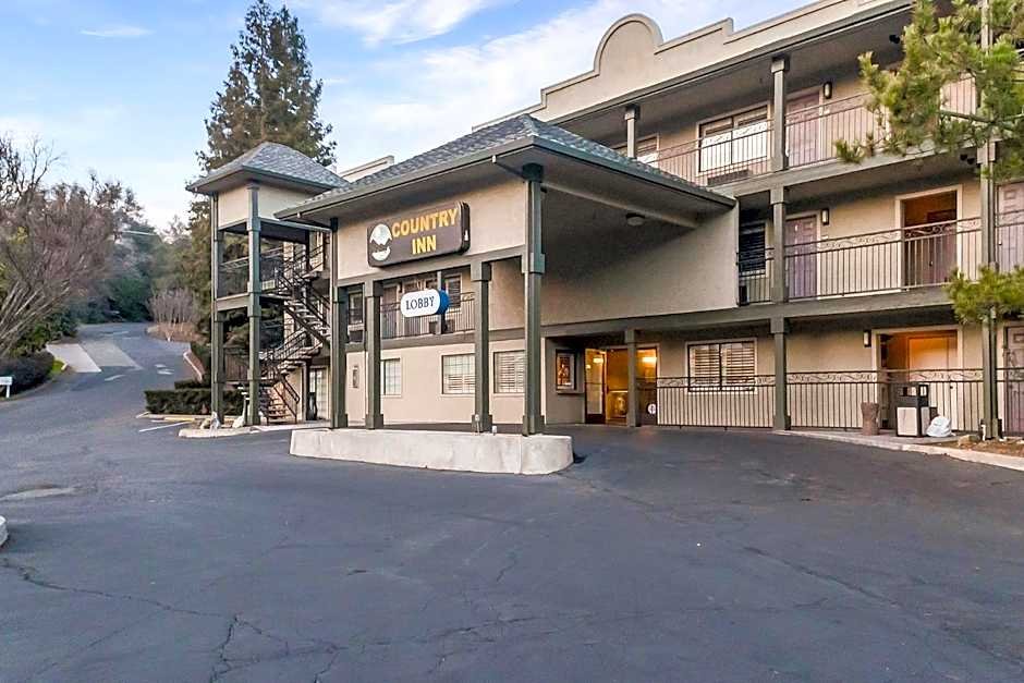 Exterior view of a two-story motel labeled 'Country Inn' with a parking lot in front and a staircase on the left side. The building has balconies and a sign indicating the lobby and room entrances.