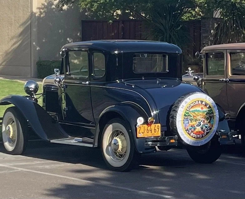 A vintage black car parked outdoors, with a California license plate and a spare tire cover featuring a colorful emblem from the Sonoara A's car club.