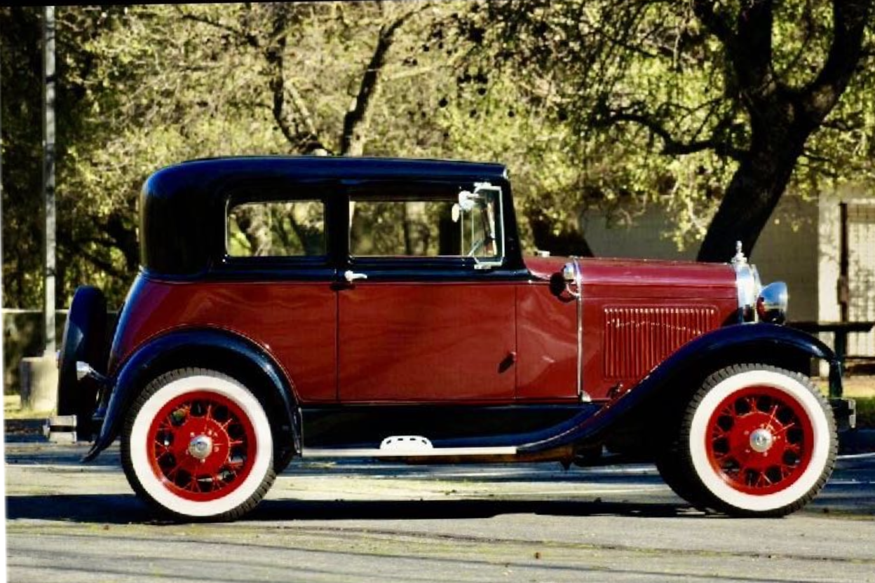 A vintage car with a black roof, red body, black fenders, and red wheels with white-wall tires parked on a street with trees in the background.