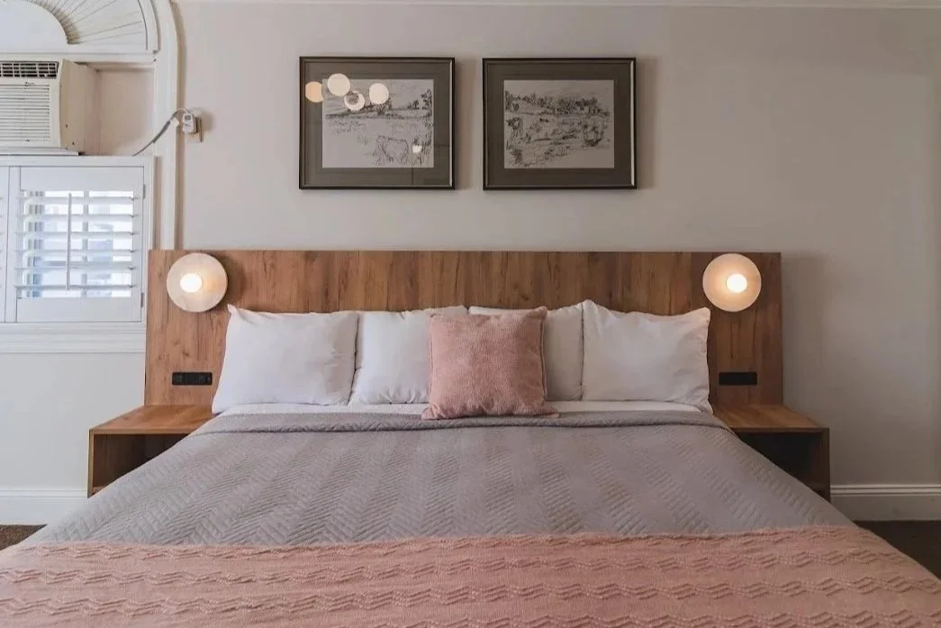 A bedroom with a bed that has a wooden headboard, three pillows, and a gray quilt. Two round wall lamps are mounted on the headboard. Above the bed are two black-framed landscape pictures. To the left is a window with white shutters.