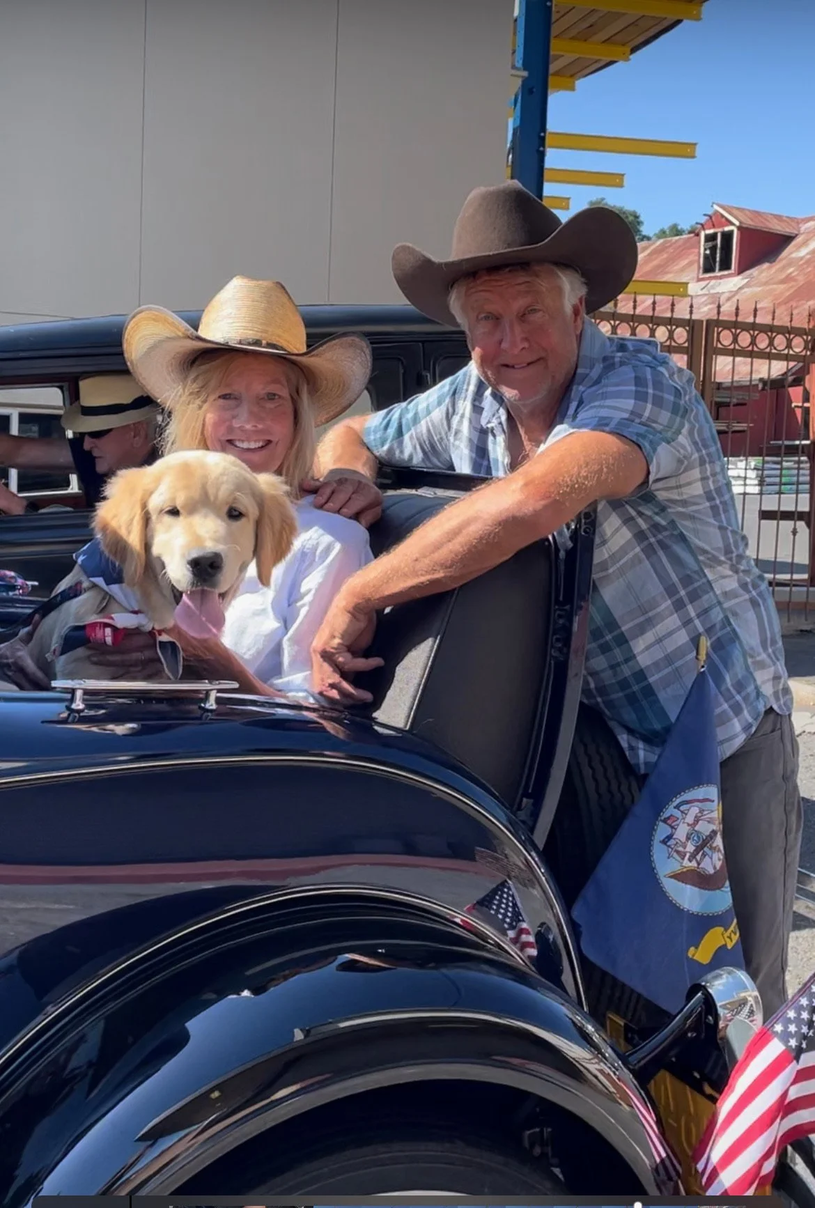 Smiling elderly man and woman with cowboy hats sit in a vintage car with a cute puppy. The man leans on the car door, and the woman is inside with the puppy, all outdoors on a sunny day.