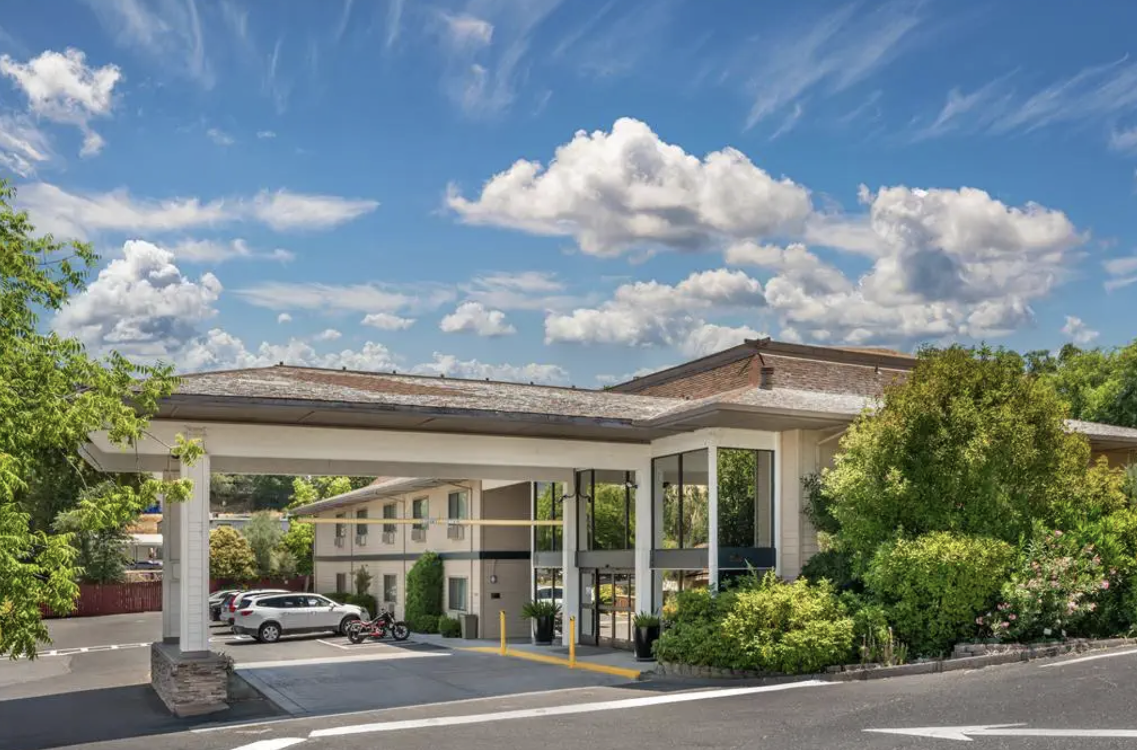 Exterior view of a modern residential or commercial building with a parking area, surrounded by greenery and under a blue sky with fluffy clouds.