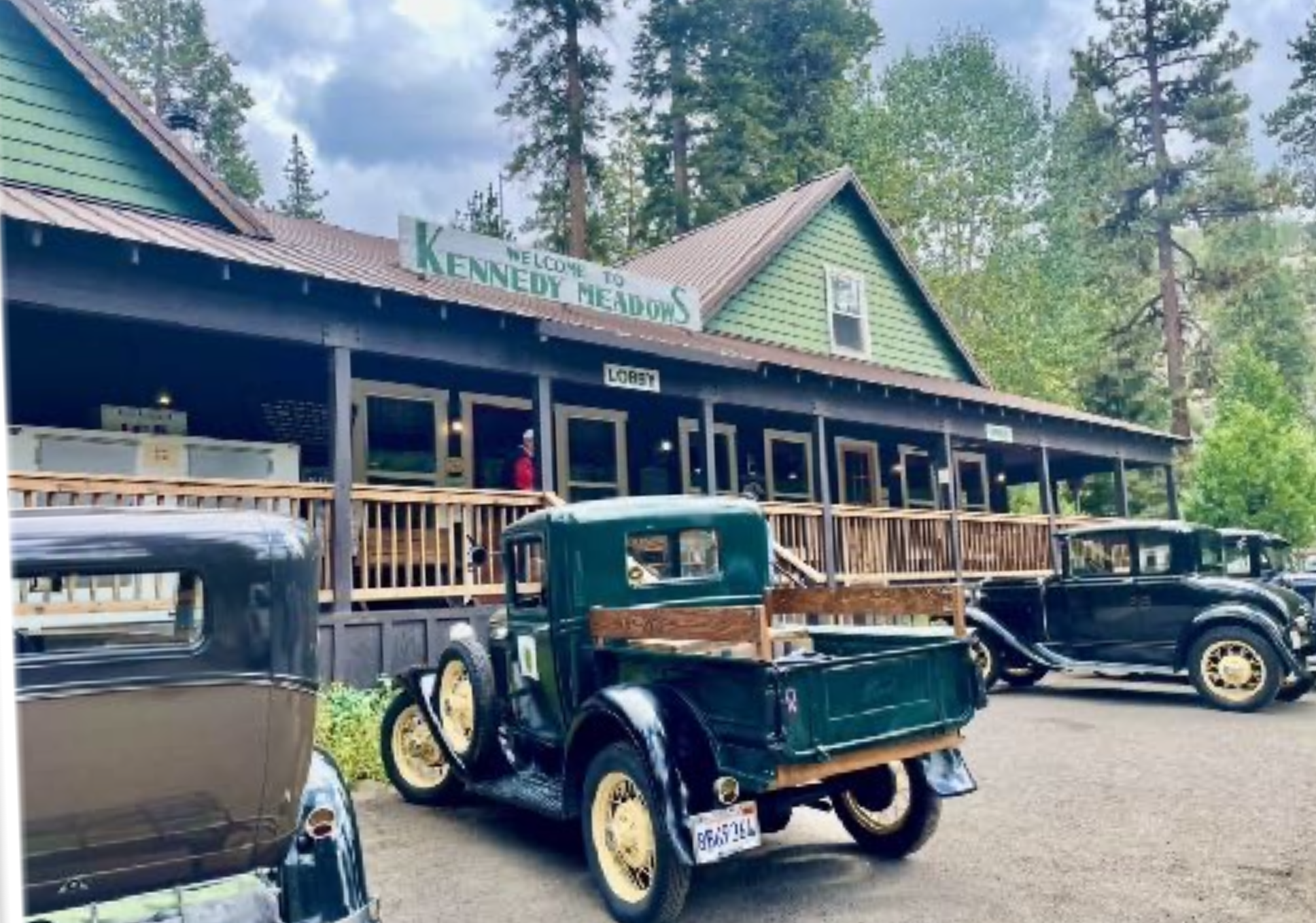 Vintage trucks parked in front of a wooden lodge with a green exterior and a sign that reads 'Welcome to Kennedy Meadows' surrounded by tall trees.
