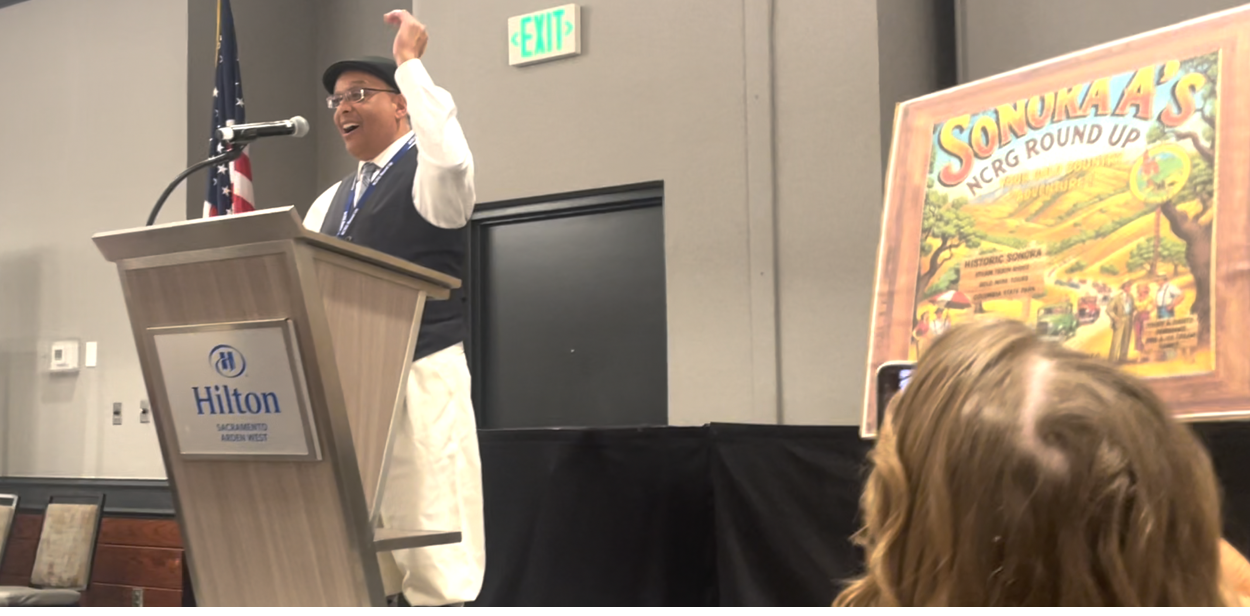 A man dressed in traditional clothing is speaking at a podium with a Hilton sign, in front of an American flag, during an event. There is a colorful poster titled 'Sonokaa's NCR Round Up' next to him, and part of an audience member's head is visible in the foreground.