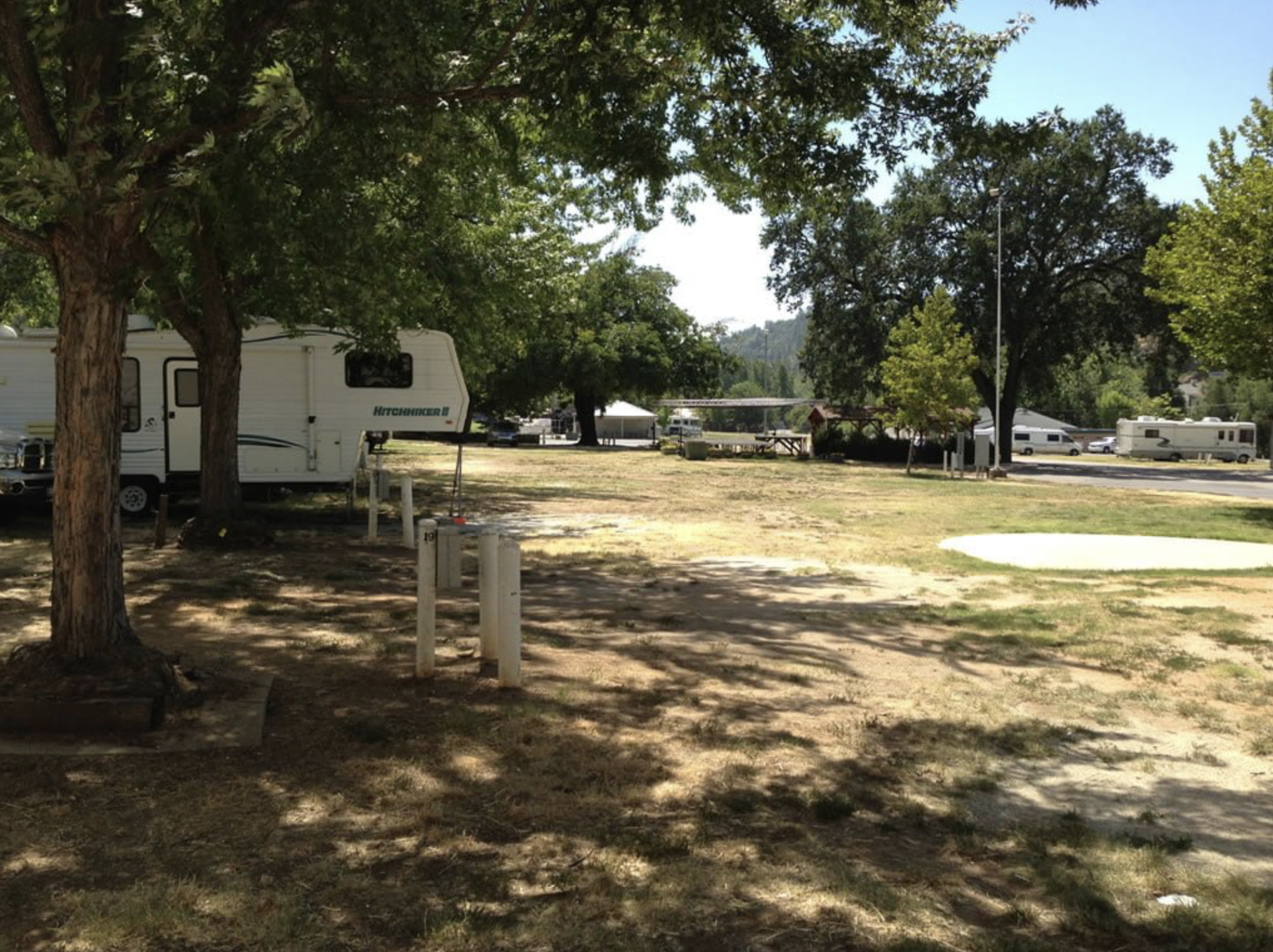 A campground with several RVs and trailers parked among trees, some picnic tables, and a sunny sky.