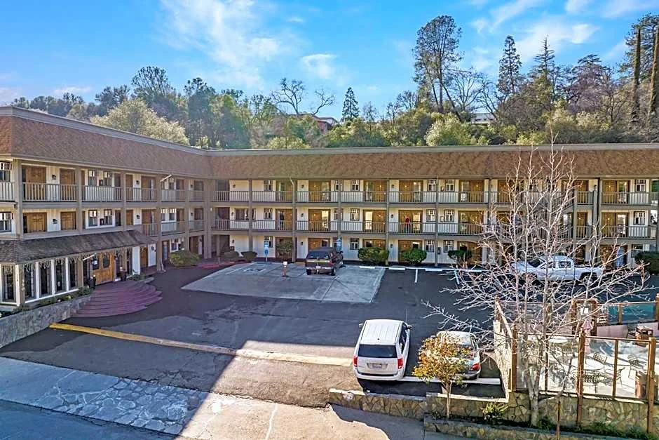 View of a three-story motel with parking lot in front, several cars parked, and trees and hills in the background under a blue sky.