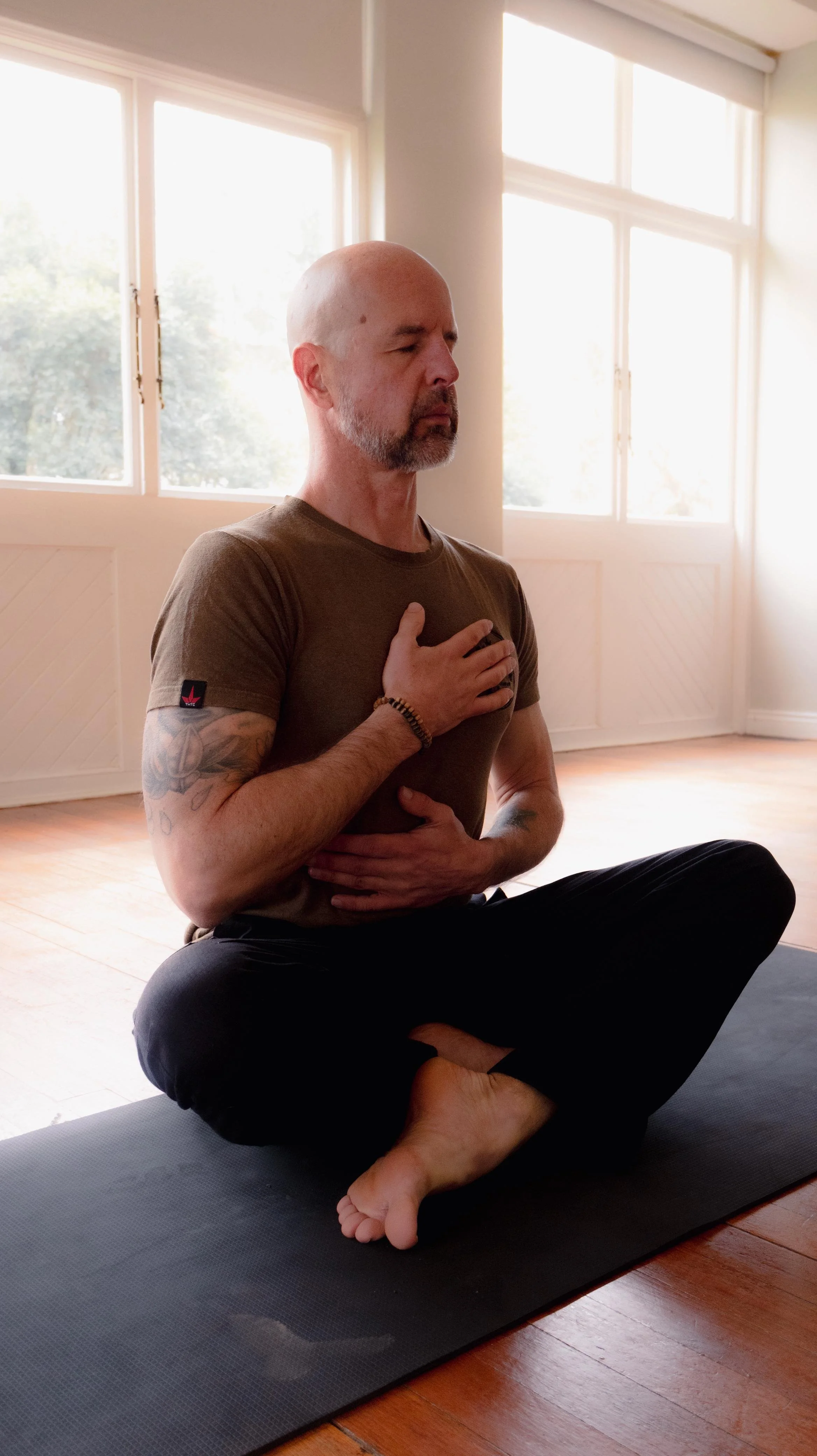A man practicing yoga indoors in a cross-legged seated position with his eyes closed and one hand on his chest and the other on his abdomen.
