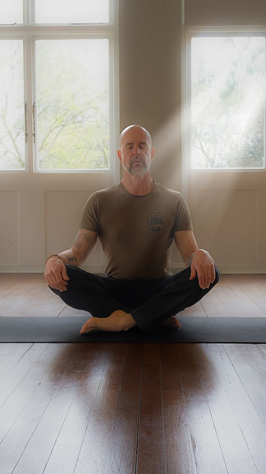 A man practicing yoga indoors, sitting cross-legged on a yoga mat with eyes closed, during daylight, with sunlight streaming through large windows behind him.