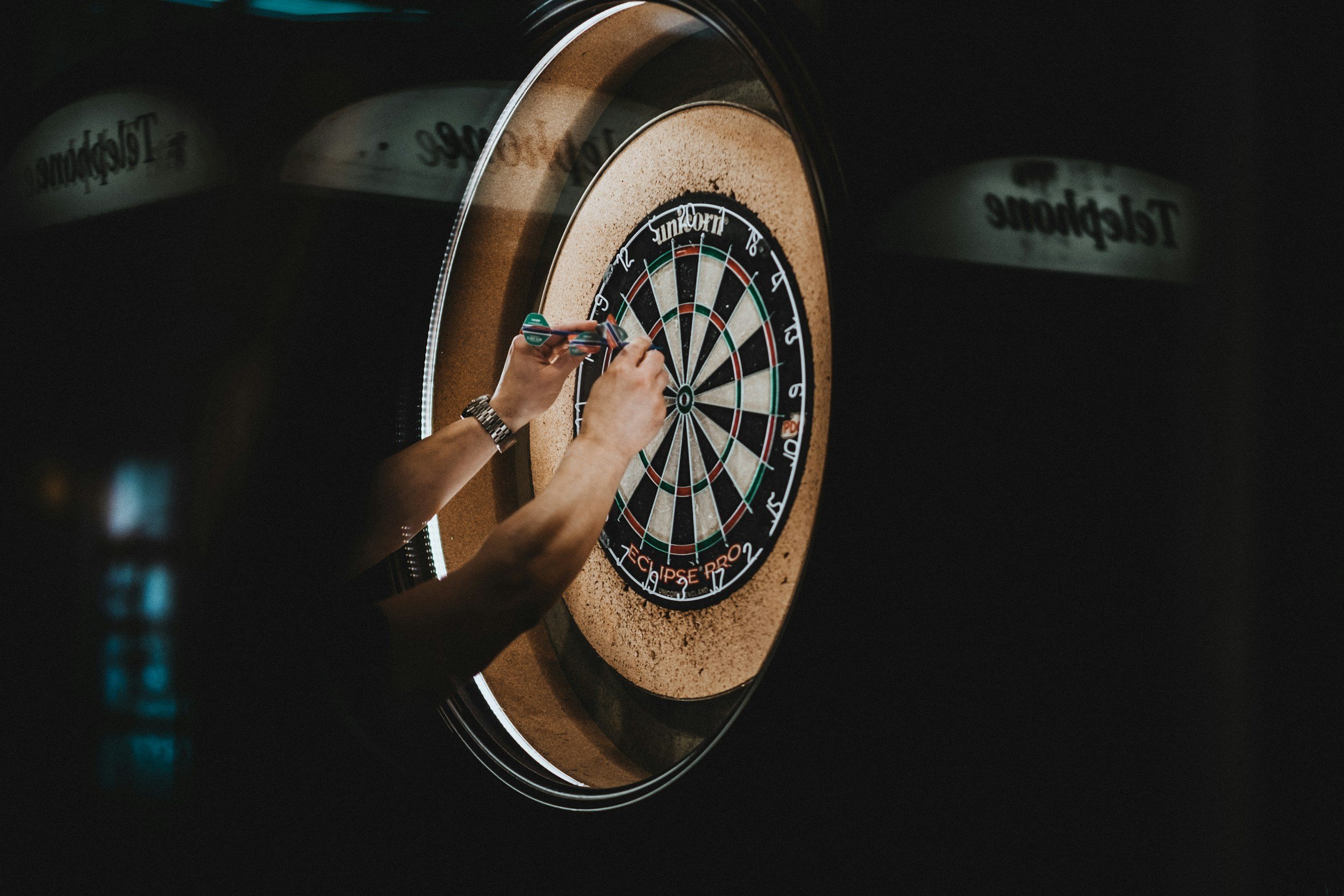 Person wirft mit einem Wurfarm auf eine Dartscheibe. Die Szene ist dunkel, die Dartscheibe ist gut beleuchtet.