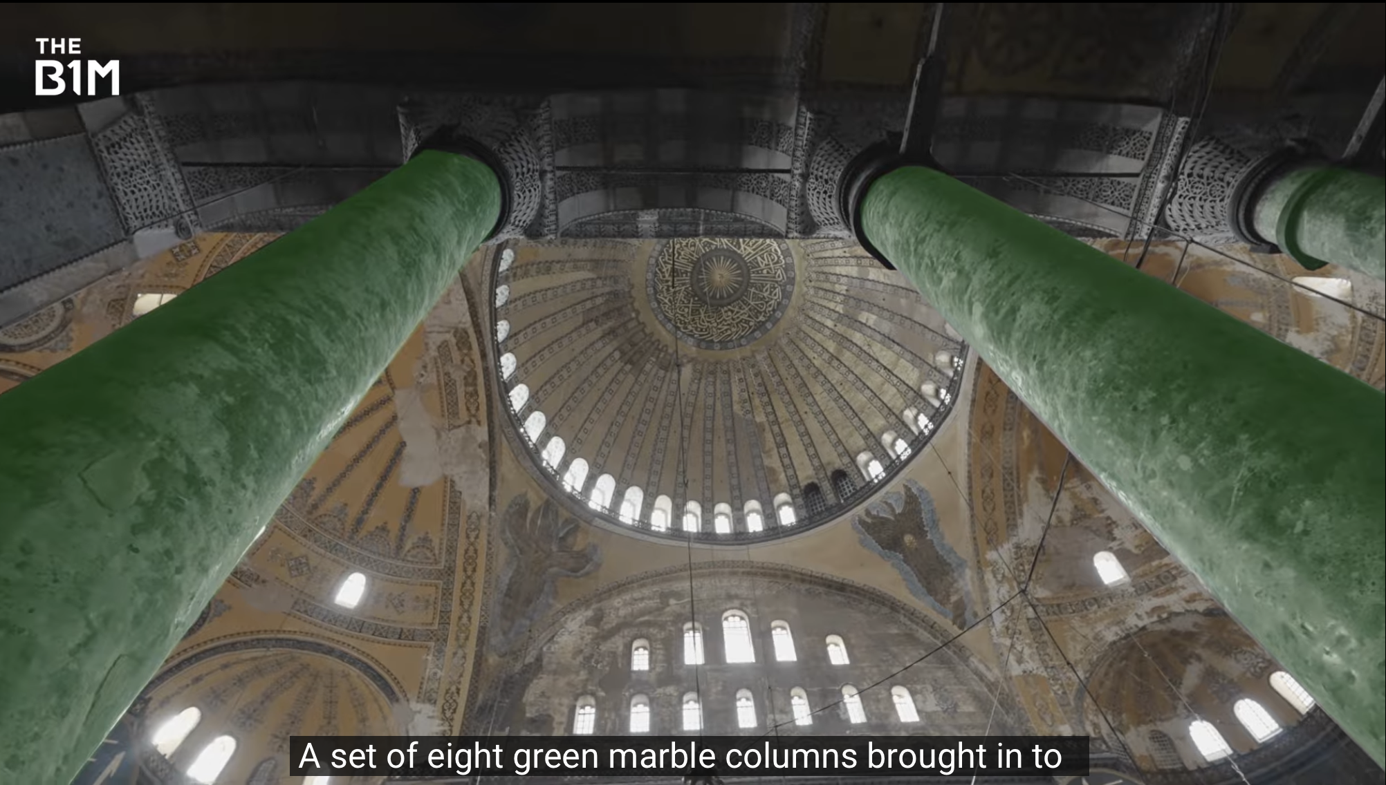 Inside view of a historic building with large green marble columns supporting a decorative ceiling with intricate patterns and a central golden chandelier.