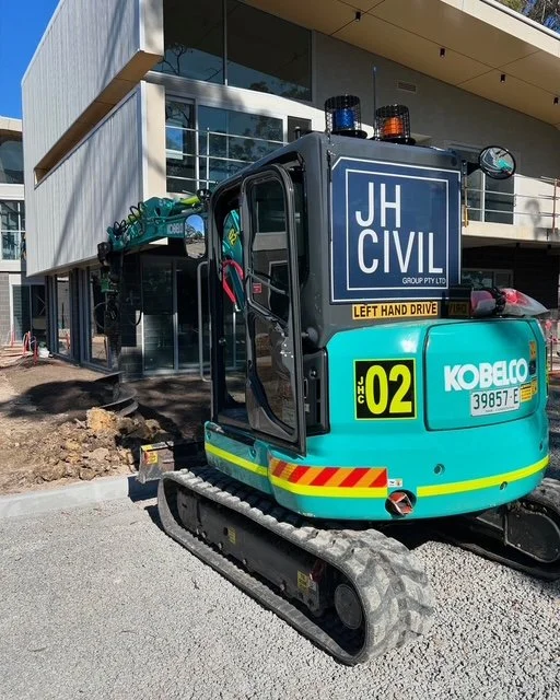 A small blue excavator parked in front of a modern glass building under construction.