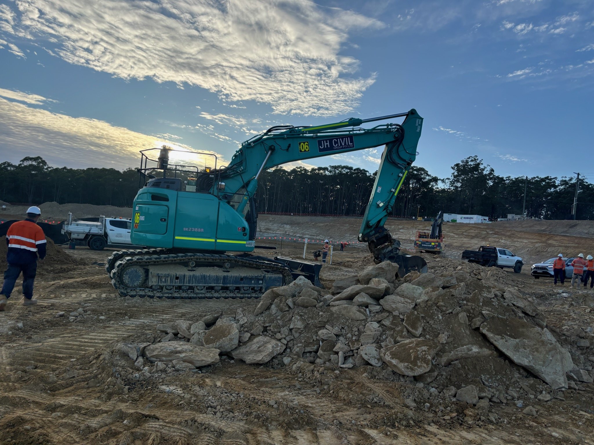 Construction site with a blue excavator moving rocks, several workers and vehicles, and a tree-lined background under a cloudy sky.