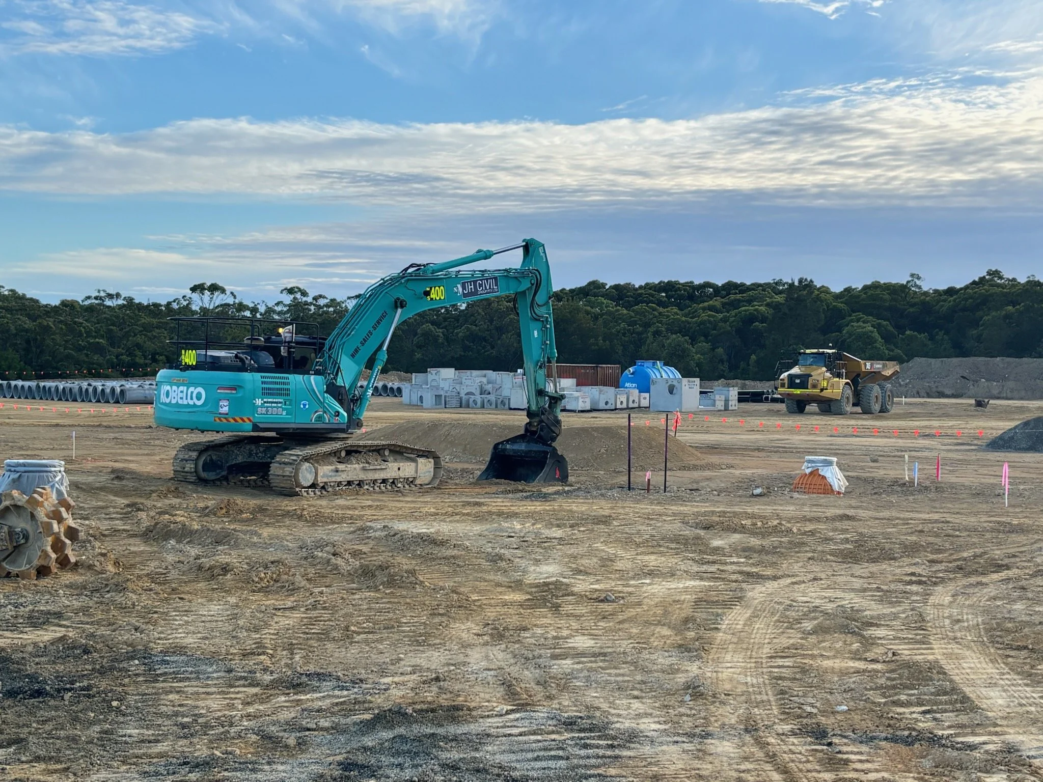 A construction site with a Komatsu excavator and a large dump truck, with items like concrete pipes and containers in the background.