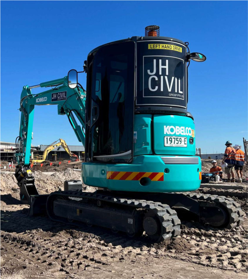 A teal Kobelco mini excavator on a construction site with a blue sky background. Construction workers are visible in the background.