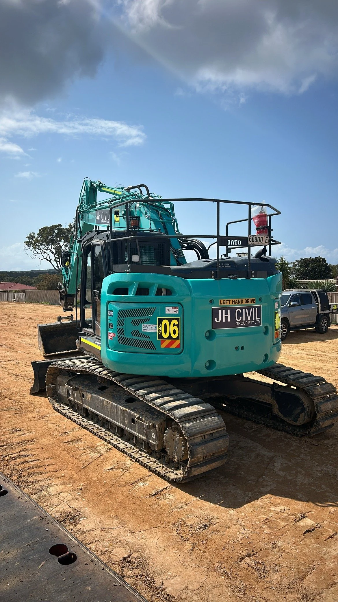 A teal-colored excavator on dirt ground with a construction site and parked vehicles in the background under a partly cloudy sky.