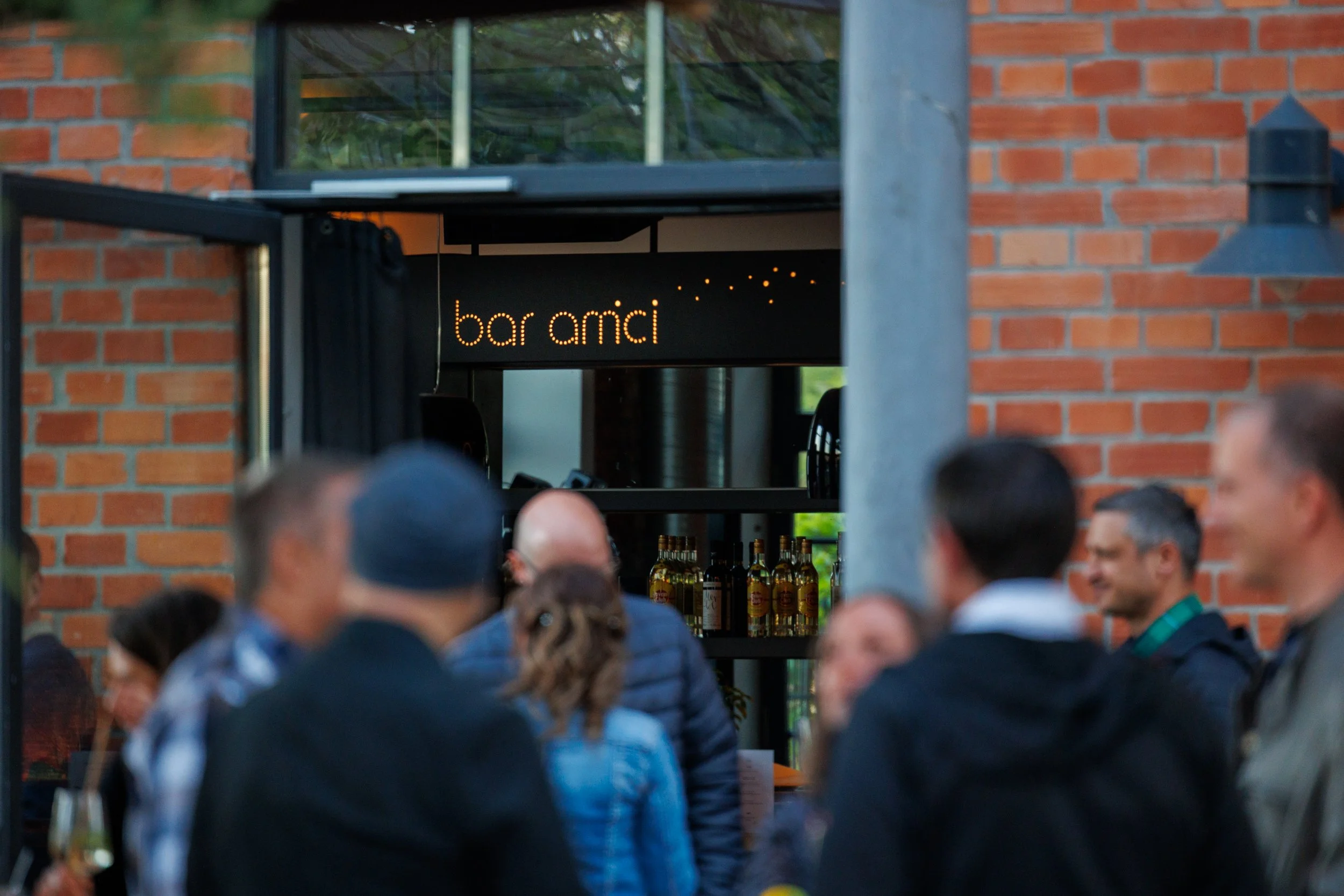 Menschen versammeln sich vor einem Restaurant oder Bar namens 'bar amici', das an einer roten Backsteinmauer mit einem großen Fenster im Hintergrund liegt. Im Inneren sind Flaschen sichtbar. Eventfotografie
